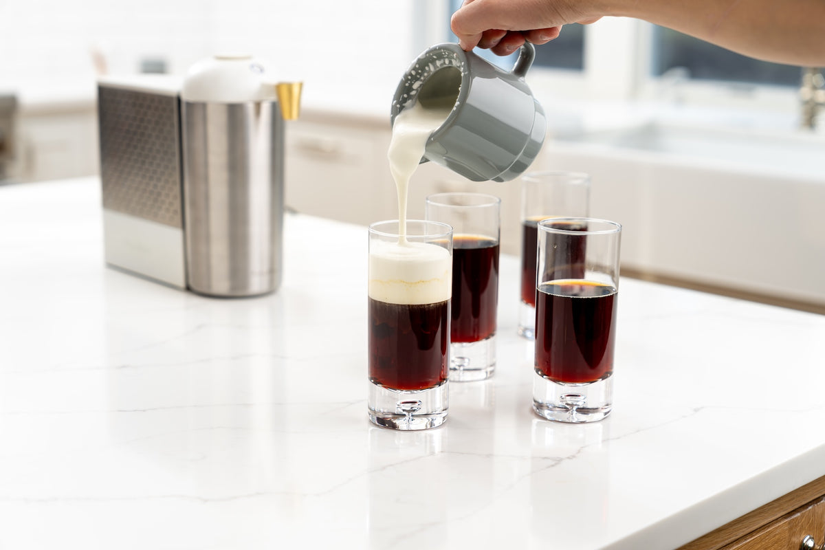 Cream pouring into a glass of cold brew beside filled coffee glasses